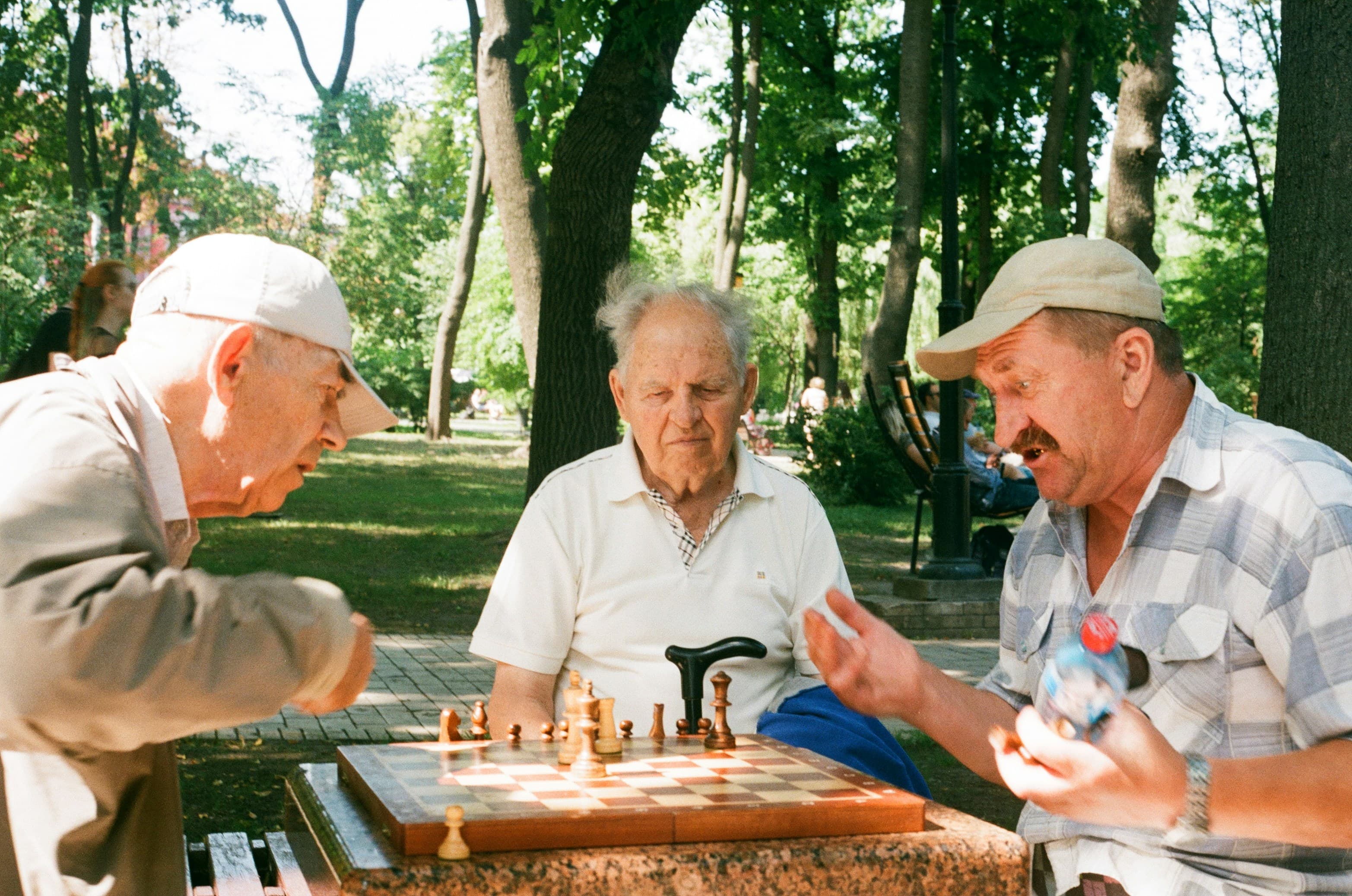 Three men gathered around a chessboard on a shaded park table mid-game