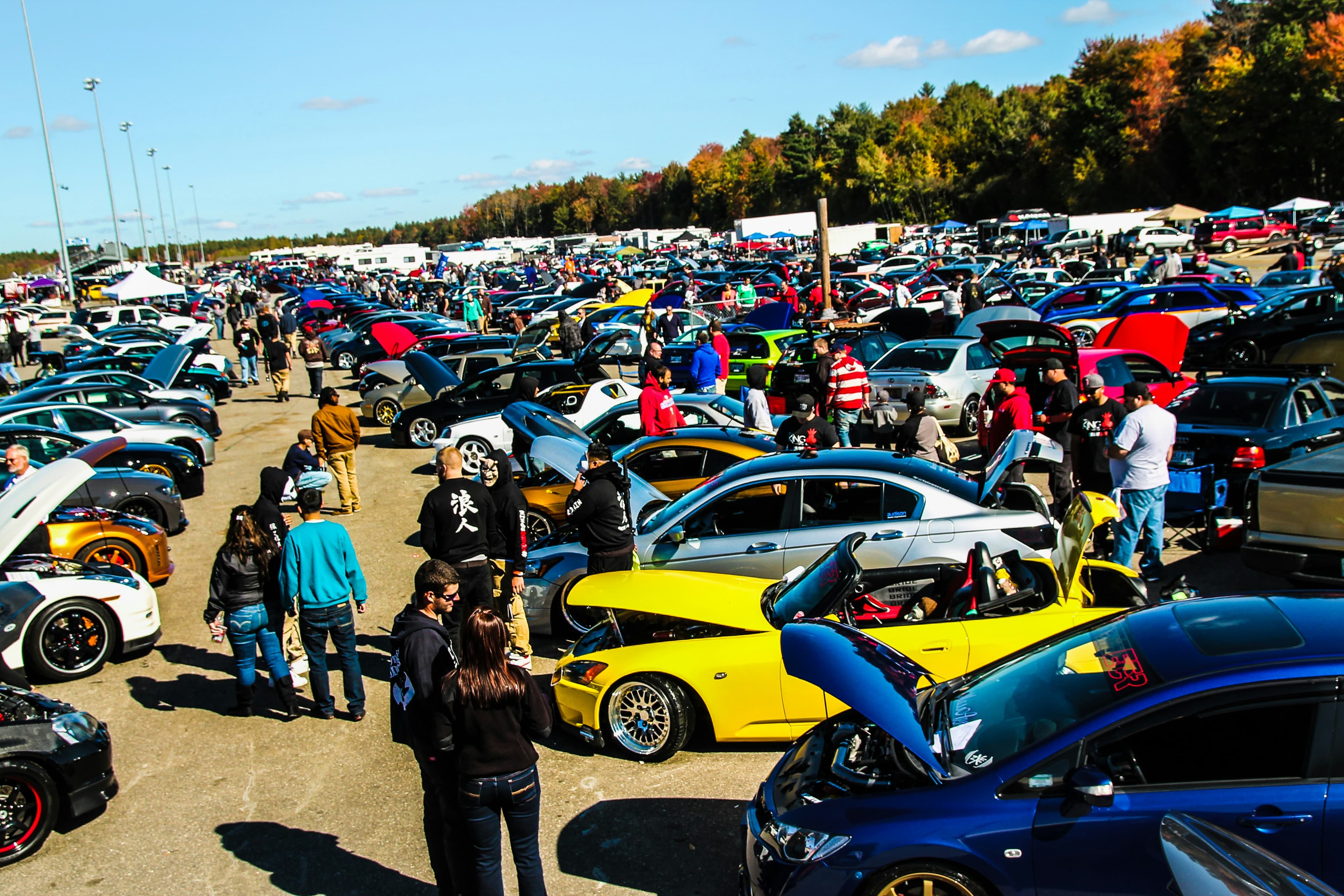 Crowd browsing rows of tuned cars with open hoods at a sunny outdoor parking lot meet