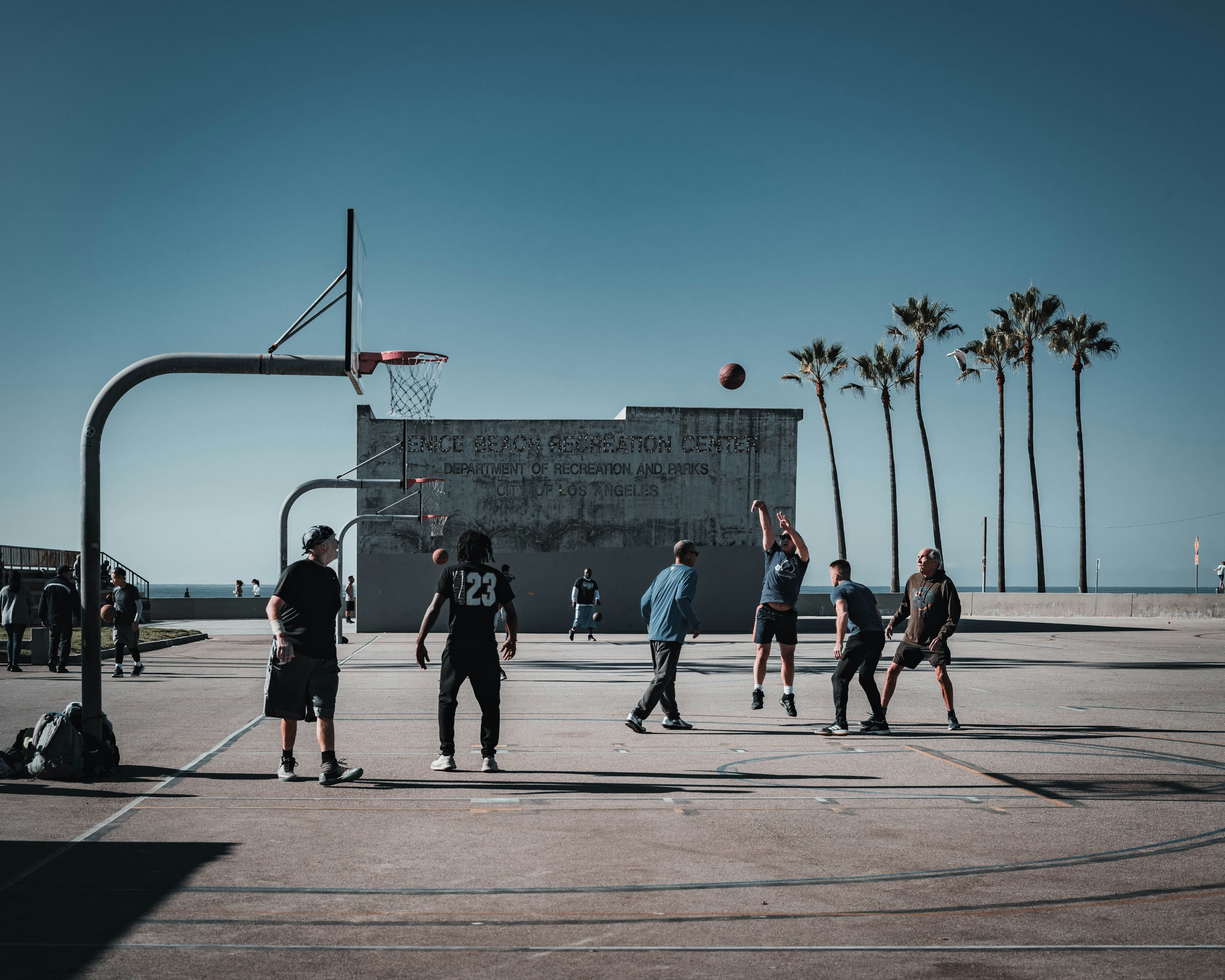 Adults playing pickup basketball on an outdoor beachside court lined with palm trees
