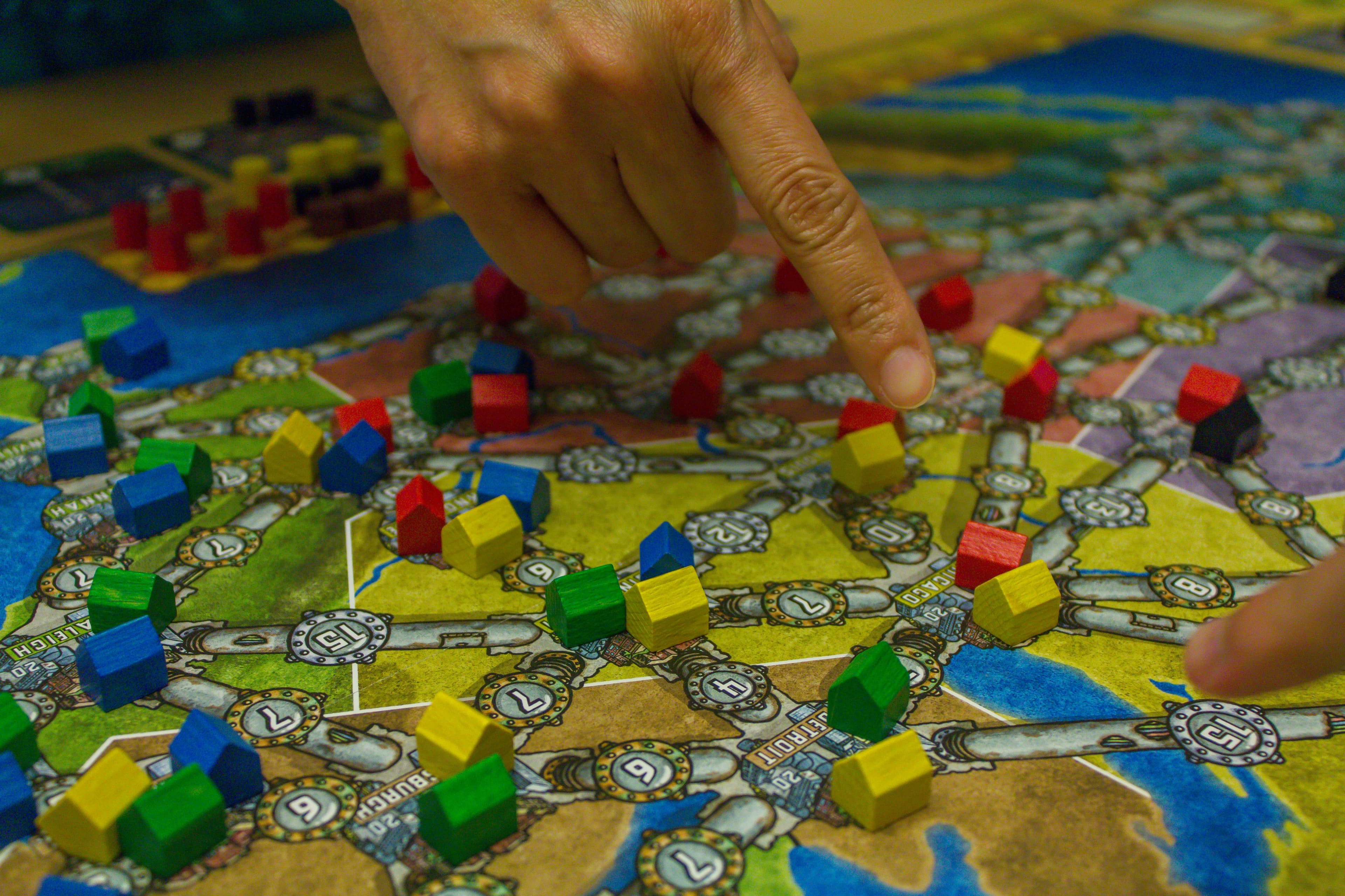 Hands pointing across a strategy board game map covered in colorful wooden pieces