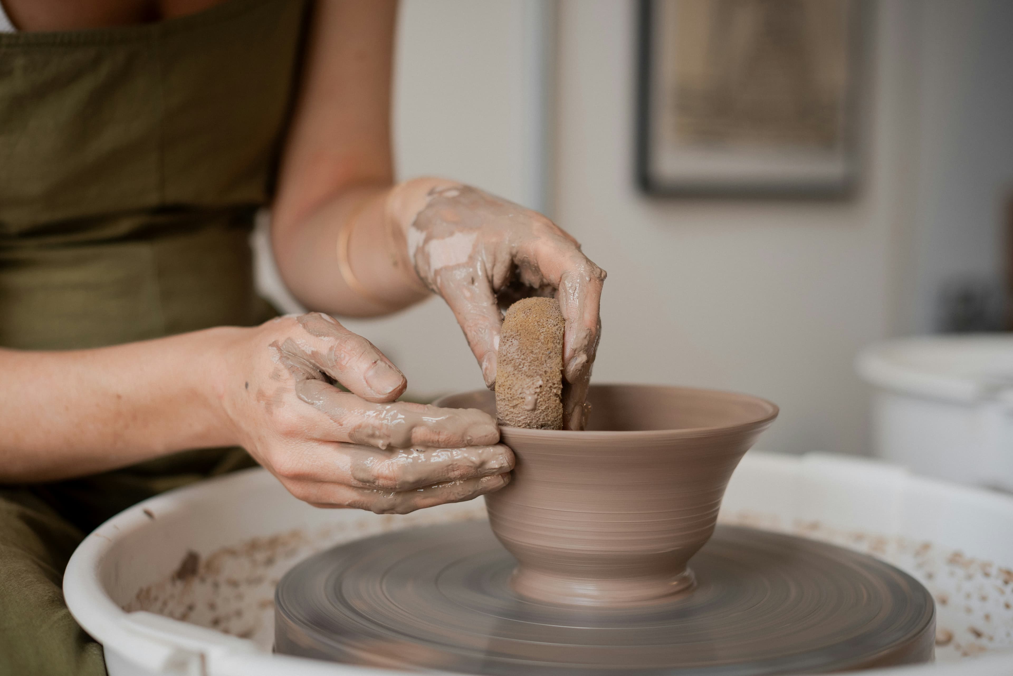 Clay-covered hands shaping a bowl with a sponge on a spinning pottery wheel